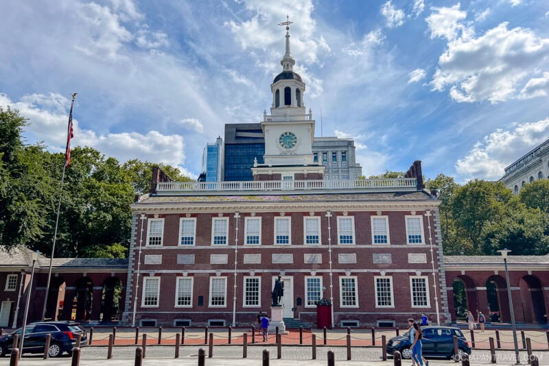 Independence Hall in Philadelphia, a historic red brick building with a central clock tower, stands under a blue sky with scattered clouds as people walk and gather in front of this iconic landmark.