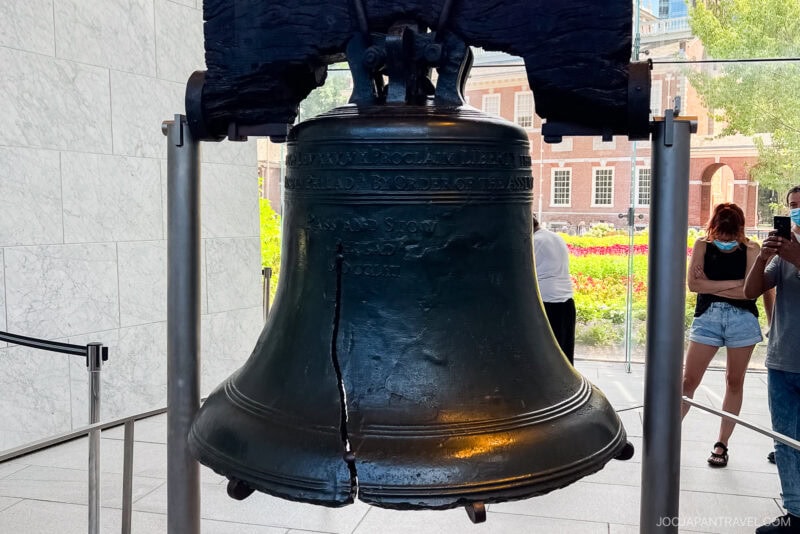 The Liberty Bell, featuring its famous crack, is displayed indoors in Philadelphia with visitors nearby. Sunlight streams through large windows, and Independence Hall is visible in the background.
