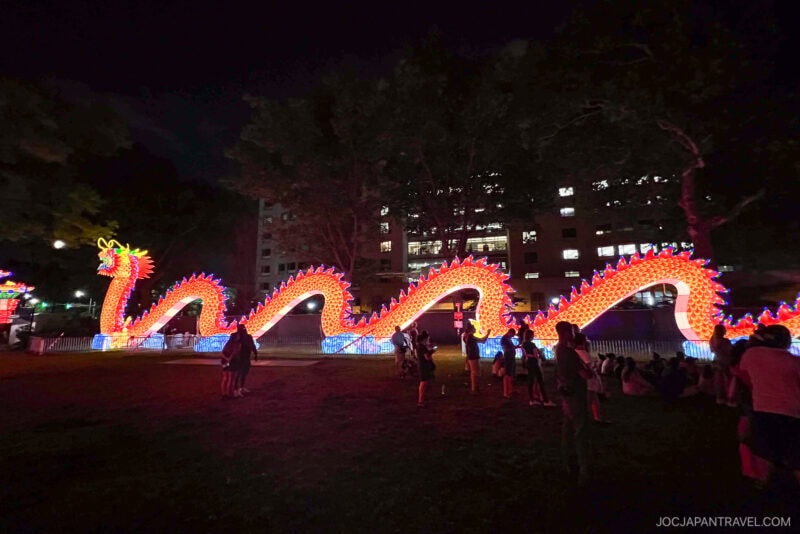 A large, brightly lit dragon lantern glows in a Philadelphia park at night, surrounded by people taking photos and admiring the display. Tall buildings and trees are visible in the background.