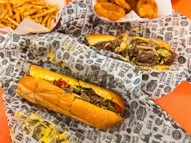 Two Philadelphia cheesesteak sandwiches on printed paper, with melted cheese, beef, and peppers. Sides of golden French fries and crispy onion rings are served in trays nearby on an orange table.