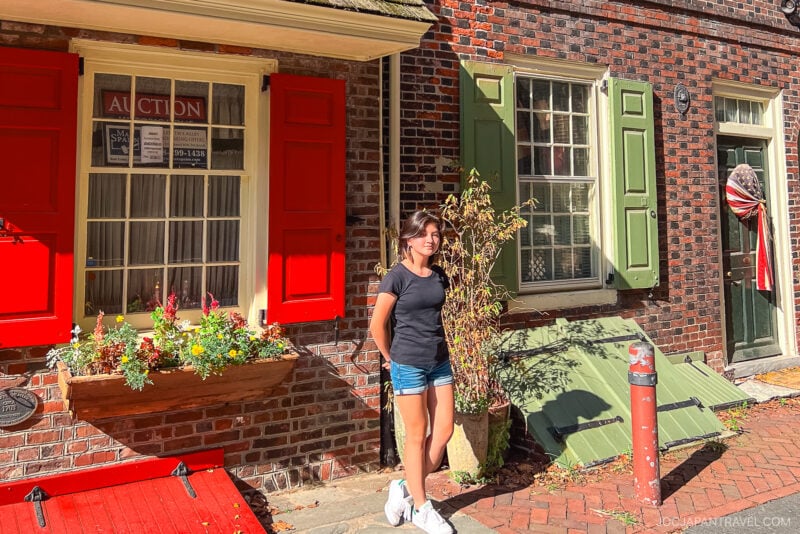 A woman in a black shirt and denim shorts stands smiling on a sunny brick sidewalk in Philadelphia, in front of a historic brick building with red and green shutters and flower boxes.