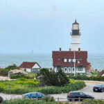 A Day in Portland, Maine – Lighthouses, Lobster Rolls and Local Flavor A tall lighthouse with a black top stands by the ocean near Portland, Maine, next to a brown-roofed building. People walk nearby and cars are parked in the foreground. The sky is overcast and another lighthouse appears in the distance.