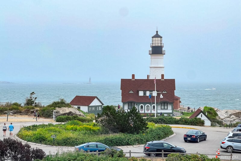 A tall lighthouse with a black top stands by the ocean near Portland, Maine, next to a brown-roofed building. People walk nearby and cars are parked in the foreground. The sky is overcast and another lighthouse appears in the distance.