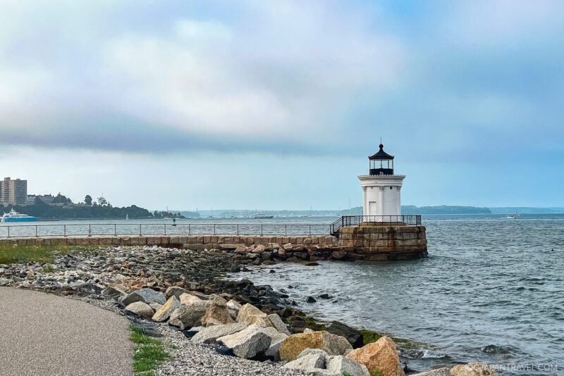 A small white lighthouse sits at the end of a stone pier by the rocky shoreline in Portland, Maine, overlooking a calm body of water under a cloudy sky. Buildings and boats are visible in the distant background.