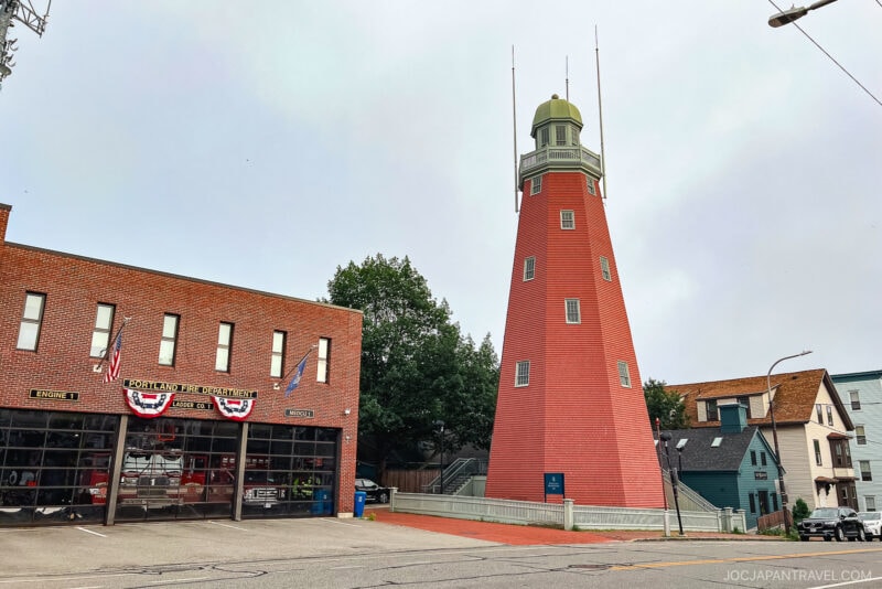 A tall, red-brick lighthouse stands beside a brick fire station in Portland, Maine, decorated with American flags and banners, with houses and trees in the background under a cloudy sky.