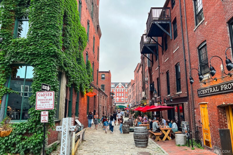 A lively cobblestone alley in Portland, Maine, lined with red brick buildings and ivy on the left, welcomes outdoor diners at wooden tables on the right. People stroll and gather under umbrellas; a sign reads Closed to vehicular traffic.