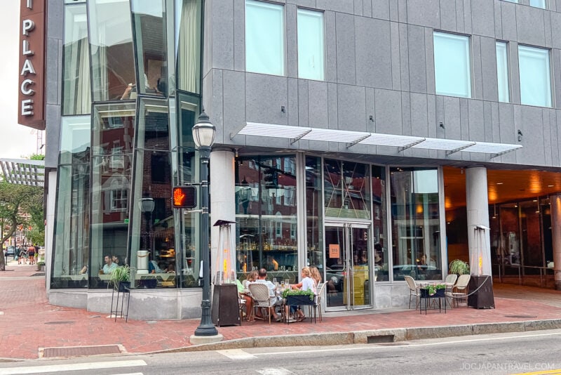 A modern city corner in Portland, Maine, with large glass windows and outdoor seating. Several people are dining at tables on the sidewalk in front of a restaurant, with a red traffic light and crosswalk visible nearby.