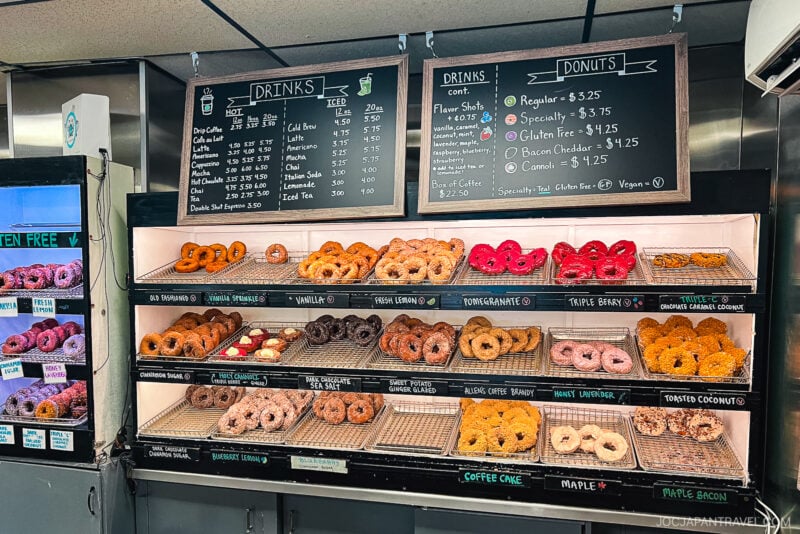A bakery display in Portland, Maine, filled with various donuts arranged on metal racks, with flavors labeled above each row. A menu board lists drink and donut prices. Some donuts are pink, chocolate, sprinkled, or covered in coconut.