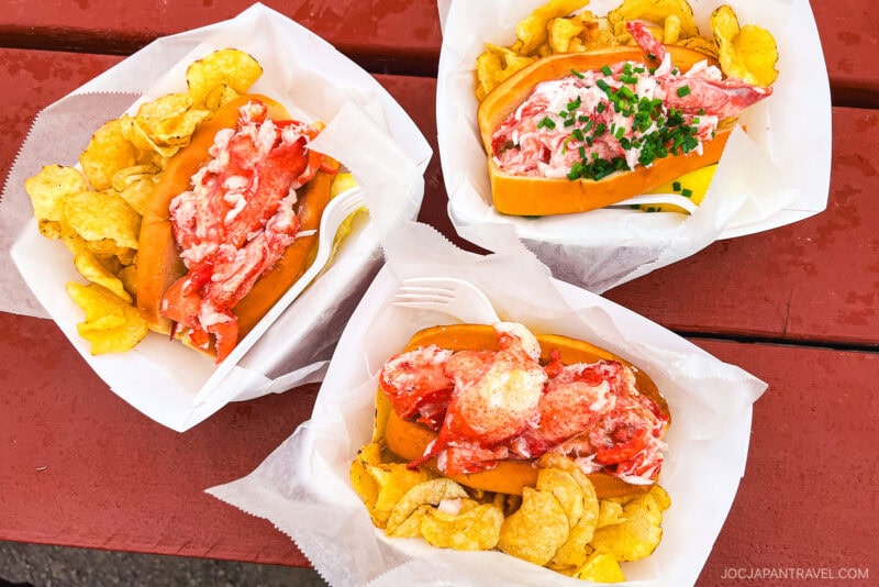Three trays of lobster rolls served with potato chips on a red picnic table in Portland, Maine. The lobster meat is piled on toasted buns, and one roll is garnished with chopped chives. A plastic fork is included with one tray.