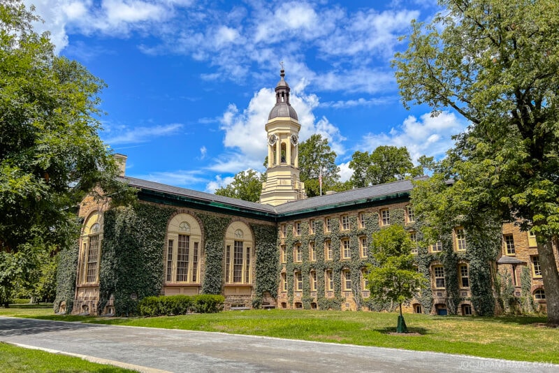 Ivy-covered stone building with tall arched windows and a central white clock tower, surrounded by green trees under a bright blue sky—an iconic scene on many Northeast College Tour stops.