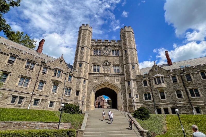 Large stone university building with a central archway, two towers, and detailed Gothic architecture. During a Northeast College Tour, people walk up wide steps toward the entrance under a blue sky with scattered clouds.