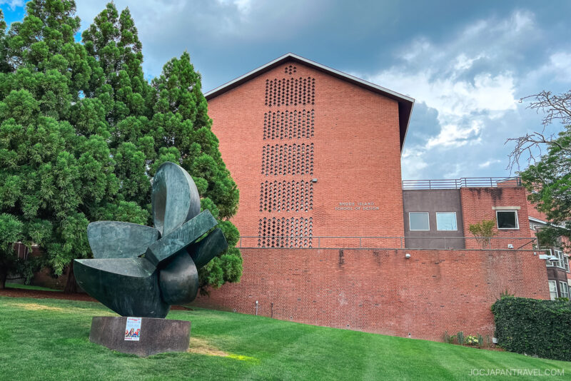A large abstract metal sculpture sits on a grassy lawn in front of a tall red brick building—an inspiring sight for any Northeast College Tour—framed by patterned windows, trees, and a partly cloudy sky.