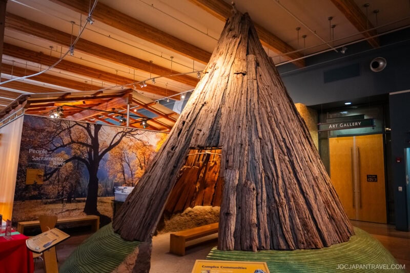 A large wooden teepee structure stands in a museum exhibit in Redding, with informational signs nearby and a photo backdrop of trees; a doorway labeled Art Gallery is visible in the background.