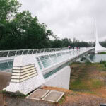 Things to Do in Redding – Nature, Parks, and Family Fun A modern white pedestrian bridge with glass panels spans over a river in Redding on a cloudy day. A few people with umbrellas cross the bridge, surrounded by greenery and trees.