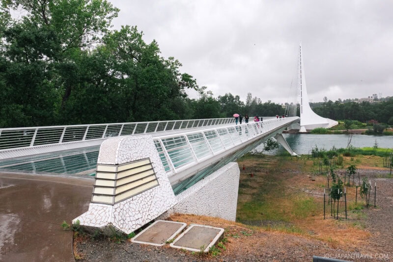 A modern white pedestrian bridge with glass panels spans over a river in Redding on a cloudy day. A few people with umbrellas cross the bridge, surrounded by greenery and trees.