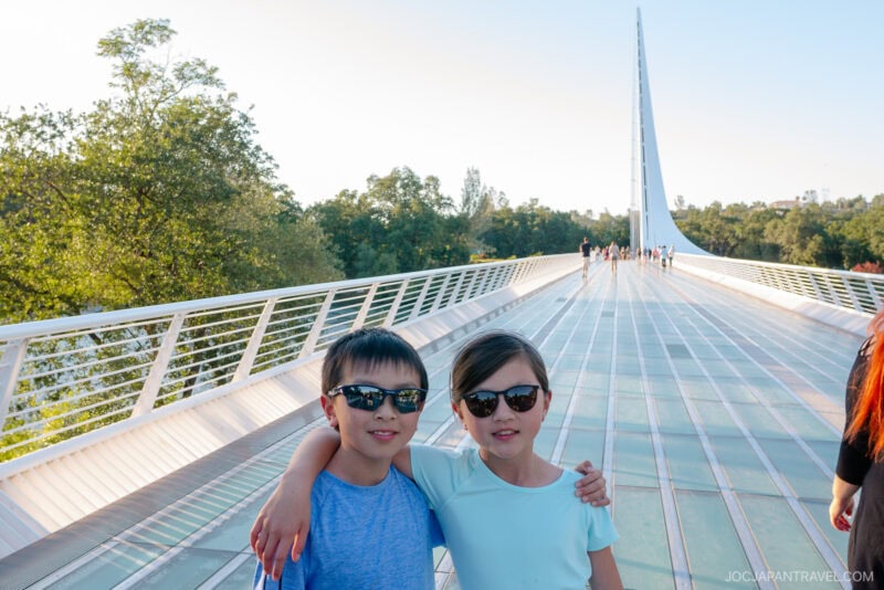 Two children wearing sunglasses stand arm-in-arm and smile on a modern white bridge in Redding, with trees and blue sky in the background. Other people are walking in the distance.