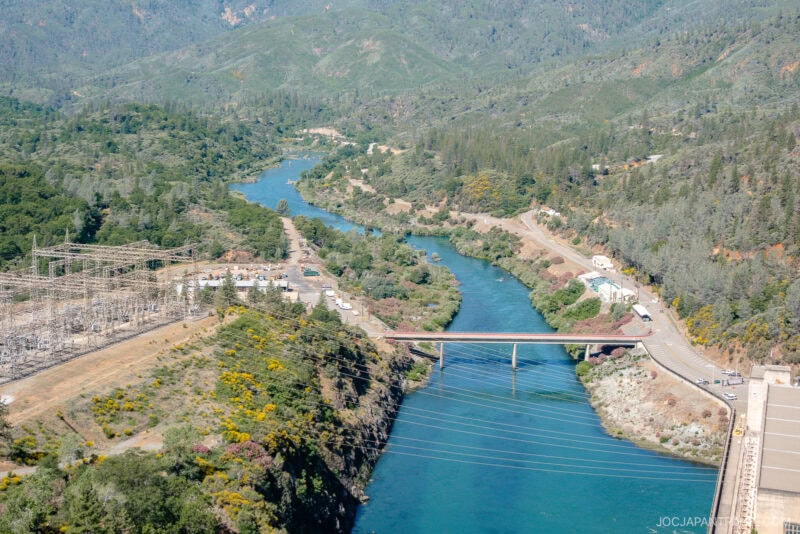Aerial view of a river with a bridge crossing over it near Redding, surrounded by green hills and forests; an electrical substation and power lines are visible on the left side.