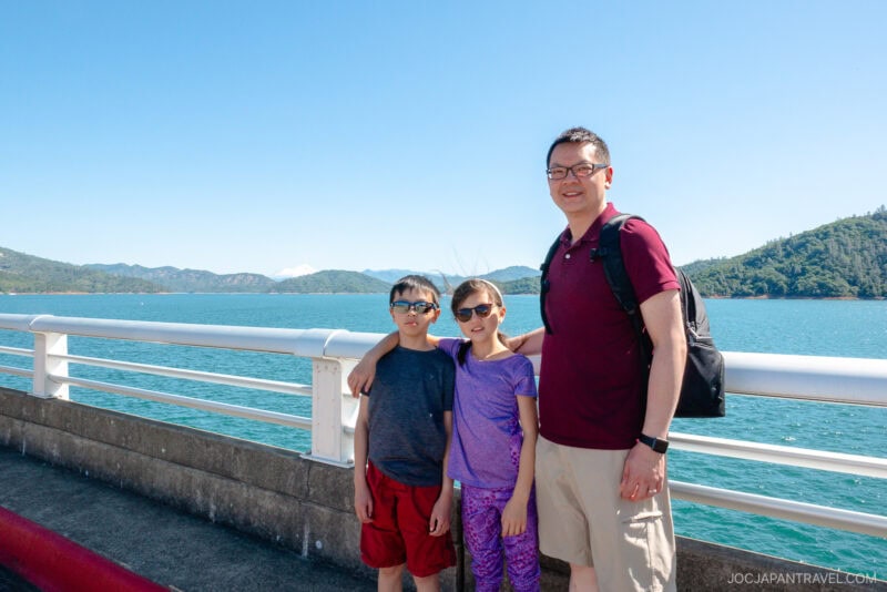 A man and two children stand on a bridge in Redding, with turquoise water and green hills in the background under a clear blue sky. They are all wearing sunglasses and smiling at the camera.