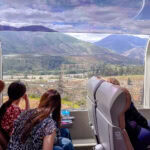 Riding the Rocky Mountaineer – Family Journey Through the Canadian Rockies Three people sit in a Rocky Mountaineer train with large curved windows, gazing out at scenic mountains, valleys, and forests beneath a partly cloudy sky—a relaxing and picturesque journey through nature.