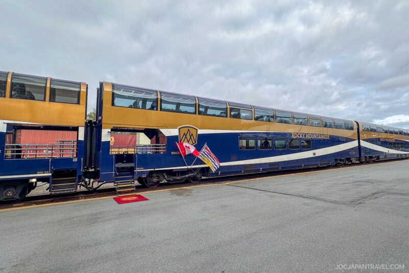 A modern, blue and gold Rocky Mountaineer double-decker train is parked at a station. Canadian and provincial flags are displayed at the entrance, with a red carpet laid out on the platform under cloudy skies.