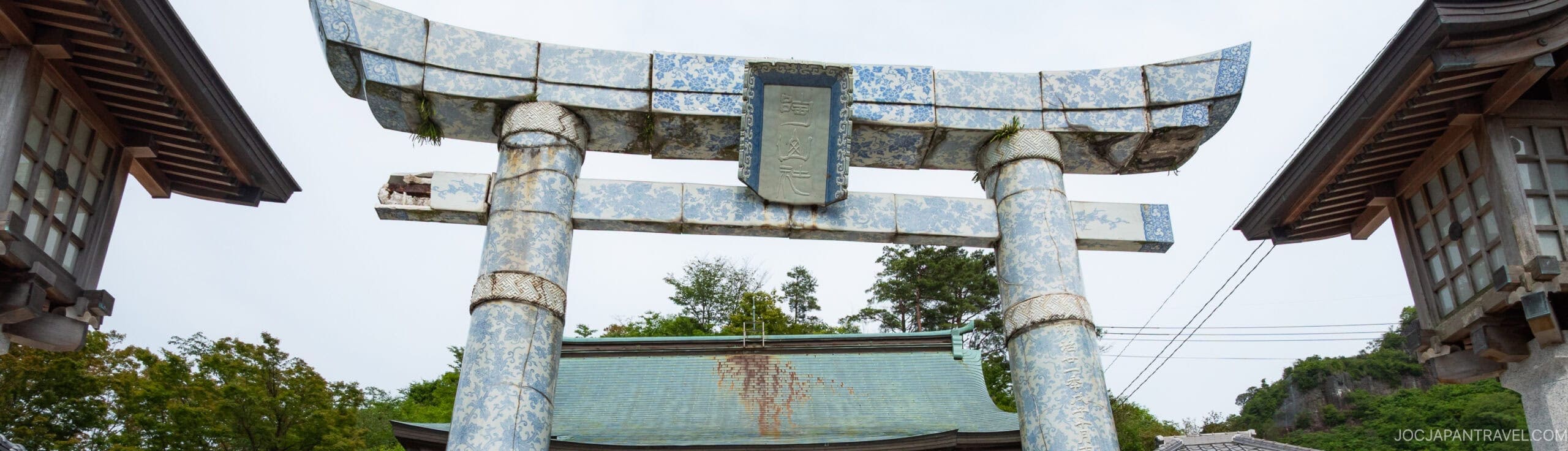 A close-up of a large, weathered torii gate with blue and white patterned tiles, standing between two traditional wooden structures at a Japanese shrine. Trees and a building roof are visible in the background.
