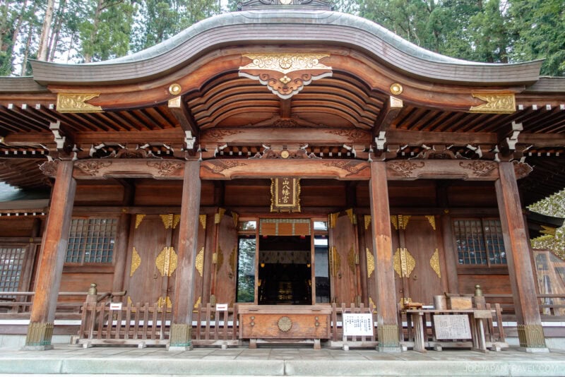 A traditional Japanese Shinto shrine, Sakurayama Hachimangu Shrine, made of wood with ornate golden decorations, a curved roof, and intricate carvings, is surrounded by tall trees in the background.