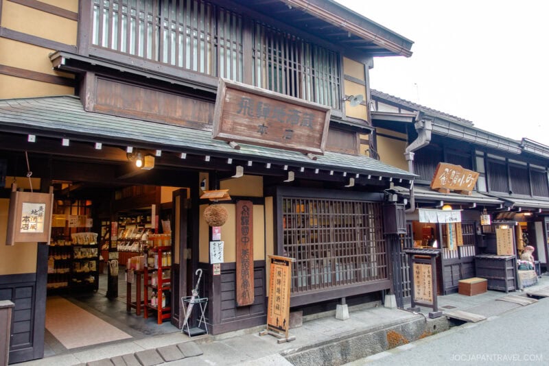 Traditional Japanese wooden shopfronts with sliding lattice doors and hanging signs line Sanmachi Suji, displaying goods inside. The narrow street outside, with a small canal and stone pavement, creates an old-town atmosphere.