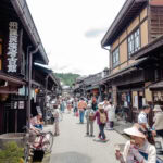 Takayama’s Old Town – Exploring Sanmachi Suji (三町筋) A lively street scene in Sanmachi Suji, a traditional Japanese town, with people walking and browsing shops housed in wooden buildings under a bright sky. Signs in Japanese hang from the buildings.