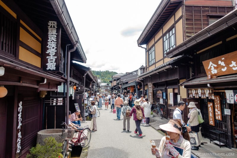 A lively street scene in Sanmachi Suji, a traditional Japanese town, with people walking and browsing shops housed in wooden buildings under a bright sky. Signs in Japanese hang from the buildings.