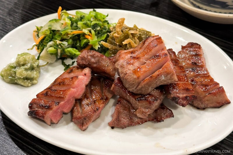 A plate of grilled beef slices with char marks, served with a side of pickled vegetables, salad, and a small portion of green paste, featured in the Sendai Food Guide on a white dish.