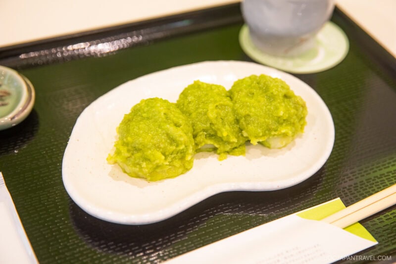 Three green, round mochi rice cakes are served on a white plate, placed on a black tray with chopsticks and a cup in the background—a classic treat featured in the Sendai Food Guide.