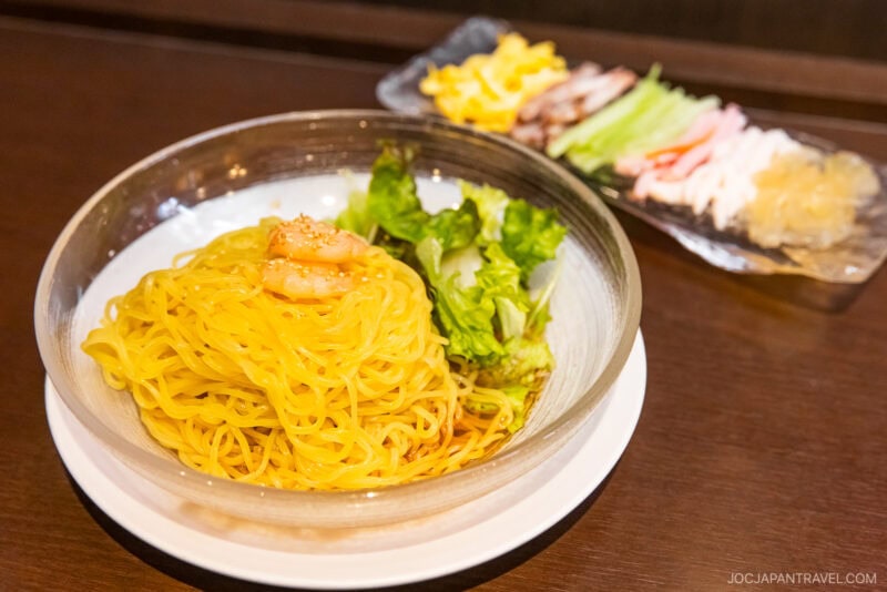 A glass bowl of cold yellow noodles topped with shrimp and lettuce sits on a white plate, with a side dish of assorted sliced vegetables and seafood blurred in the background—a fresh highlight from the Sendai Food Guide.