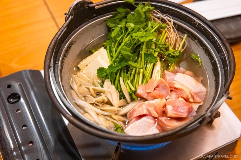 A Japanese hot pot featured in the Sendai Food Guide, with raw chicken, tofu, mushrooms, and fresh greens simmering in broth on a portable stovetop atop a wooden table.