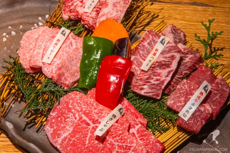 A platter of assorted raw Wagyu beef cuts, labeled with tags and garnished with green leaves, sits alongside sliced bell peppers and carrot on a rustic tray—a true highlight for any Sendai Food Guide.