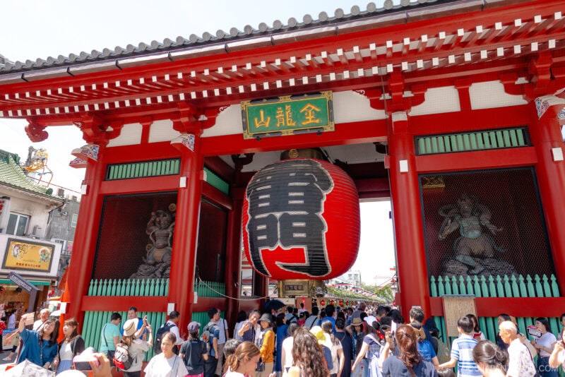 A large red lantern hangs at the entrance of Tokyo&rsquo;s Senso-ji Temple, framed by bold red columns and a crowd of visitors below. Statues are visible in alcoves on either side of the gate.