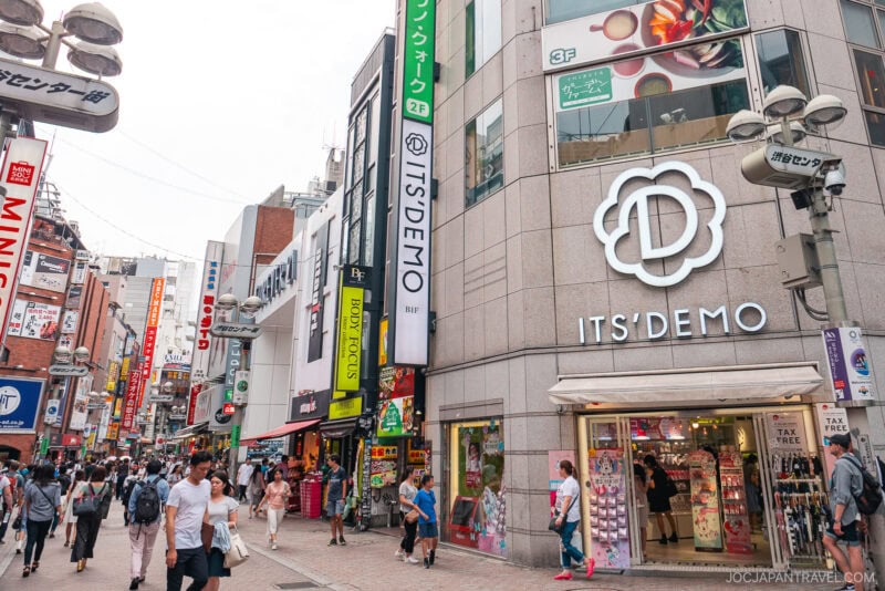 A busy street in Japan&rsquo;s Shibuya Center Gai with people walking, colorful shop signs, and a large &ldquo;IT&rsquo;S DEMO&rdquo; store on the corner. The lively atmosphere is filled with various shops and vibrant advertisements.