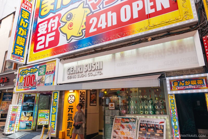 A person walks past the brightly lit entrance of GENKI SUSHI in Japan's lively Shibuya Center Gai, surrounded by colorful signs and advertisements, including a large 24h OPEN sign above the storefront.