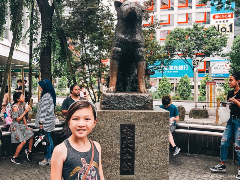 A young girl smiles in front of the famous Hachiko dog statue in Shibuya, Tokyo, Japan. People and city buildings fill the background, creating a lively urban atmosphere.