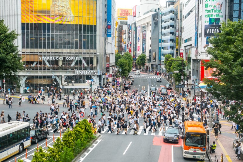 A busy crosswalk in Shibuya, Tokyo, with crowds of people crossing the street in multiple directions, surrounded by tall buildings, shops, bright signs, and vehicles. Experience the iconic energy of Shibuya&rsquo;s bustling cityscape.