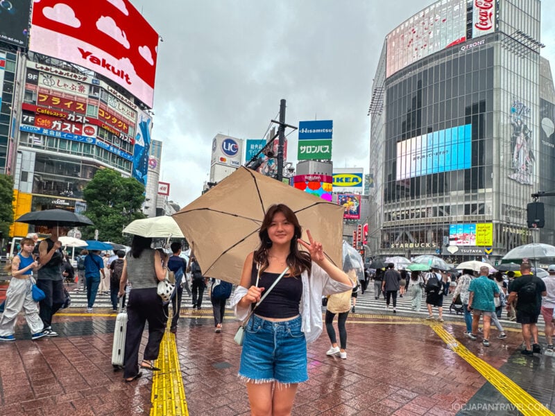 A young woman holding an umbrella and flashing a peace sign stands at Shibuya Crossing in Tokyo on a rainy day, surrounded by people, billboards, and vibrant city lights.
