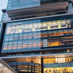 The modern multi-story Shibuya Hikarie features large glass windows, a brightly lit interior, and people visible inside. The building boasts a sleek, contemporary design in the heart of the city.