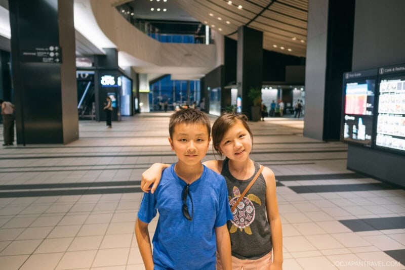 Two children stand close together indoors at Shibuya Hikarie, smiling at the camera. The boy wears a blue shirt and the girl a gray dress. The spacious, modern interior features tiled floors, high ceilings, and people in the background.