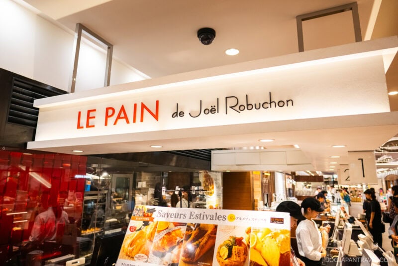 Bakery storefront at Shibuya Hikarie with a sign reading “LE PAIN de Joël Robuchon” above the counter, and a display of food advertisements and people ordering inside a busy, well-lit shop.