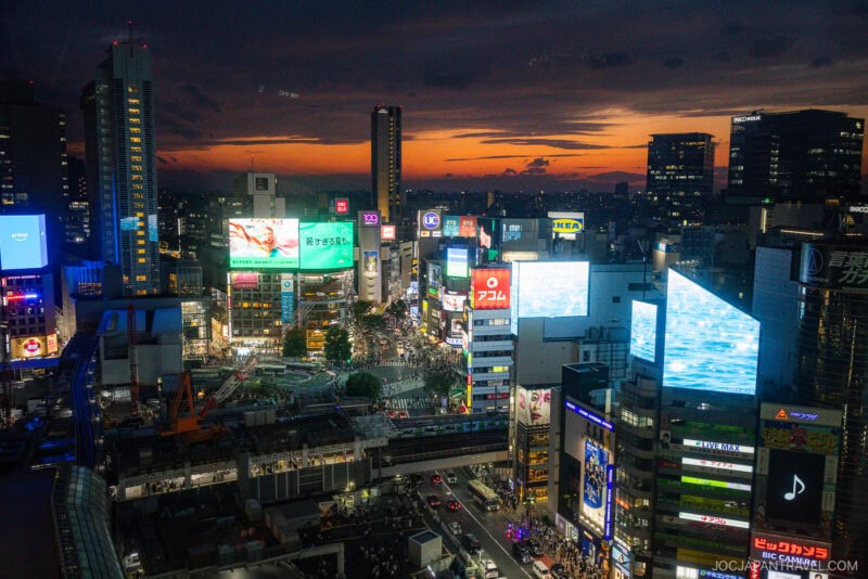 A vibrant cityscape of Shibuya in Tokyo at sunset, featuring the iconic Shibuya Hikarie among illuminated billboards, bustling streets, and tall buildings under a dramatic orange and blue sky.