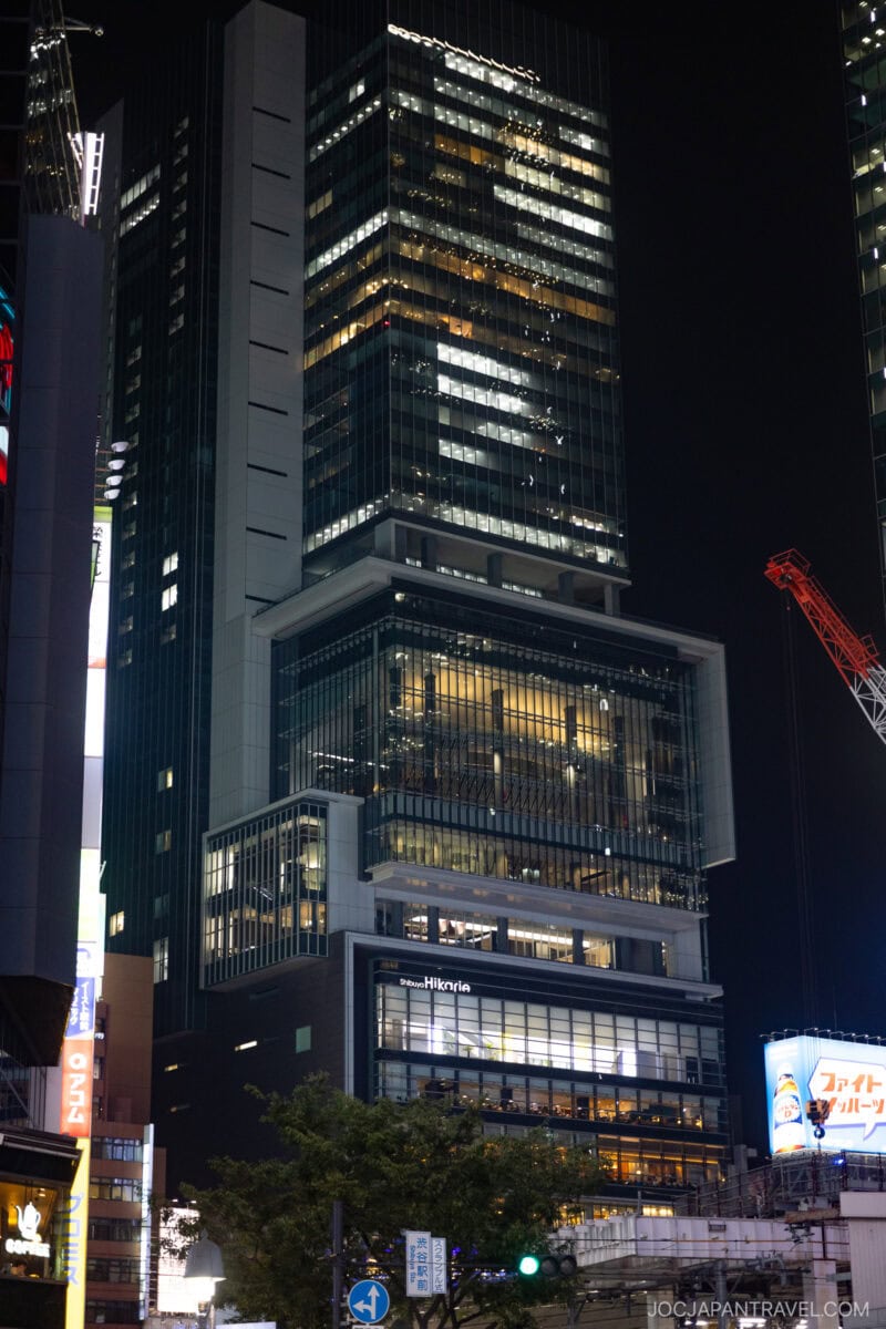 A tall, modern skyscraper lit up at night in an urban area, with the word Shibuya Hikarie illuminated on one of its lower floors and other high-rise buildings and neon signs visible nearby.