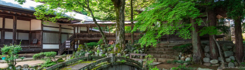 A serene Japanese garden with lush green trees, a small stone bridge, and traditional wooden buildings surrounding a peaceful courtyard. Sunlight filters through the foliage, highlighting the tranquil setting.