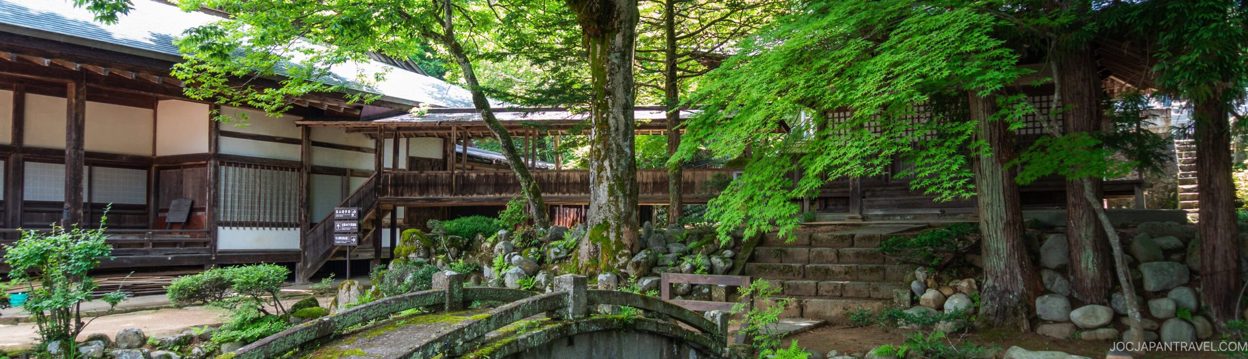 A serene Japanese garden with lush green trees, a small stone bridge, and traditional wooden buildings surrounding a peaceful courtyard. Sunlight filters through the foliage, highlighting the tranquil setting.