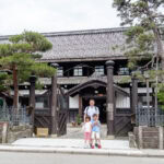 Takayama City Archives Museum (高山市政記念館) – Unveiling the Past A woman and two children stand smiling in front of the Takayama City Archives Museum, a traditional Japanese wooden building with a tiled roof, surrounded by trees and a gated entrance on a cloudy day.