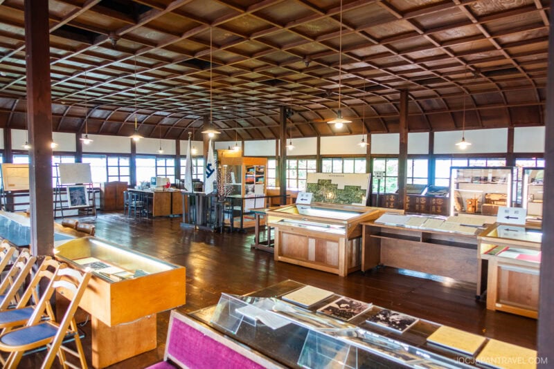 A spacious room with wooden floors and a lattice ceiling at the Takayama City Archives Museum, displaying historical documents, photographs, and artifacts in glass cases and on tables, with chairs arranged along one side.