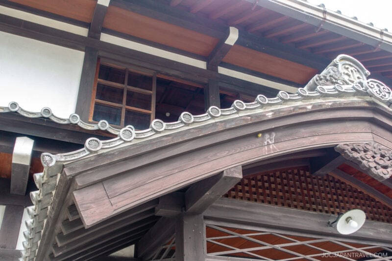 Traditional Japanese wooden building with ornate, curved roof tiles, intricate woodwork, and lattice designs under the eaves—features characteristic of the Takayama City Archives Museum—viewed from below.